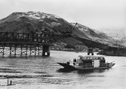 This picture shows a meeting of old and new transport generation in Sogndal. In the background the Loftesnes bridge is under construction to be opened in 1958. In the foreground we see the ferry used for transporting cars across the strait. The ferry pilot was Bernt Pedersen.