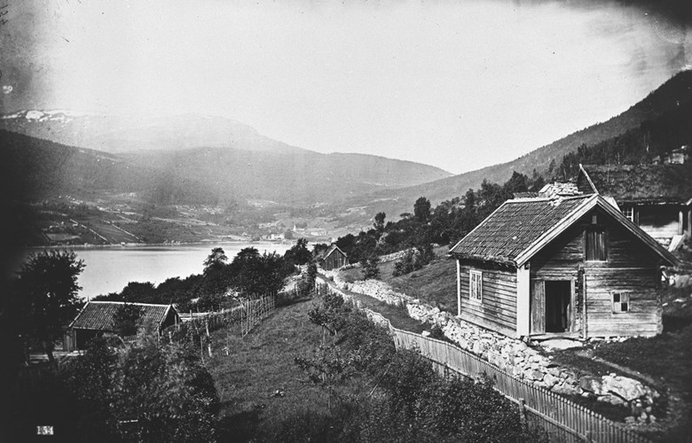The picture dates from 1880 and shows the old cart road built in 1848. The picture is taken from upper Amla towards the Kaupanger estate and the stave church in the centre of the picture. The houses on the picture are located where the first buildings of the Sogn Folkemuseum were put up about 1900. 