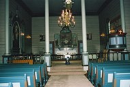 From the interior of the nave seen towards the chancel. The main colour scheme is grey and blue. The walls and the ceilings in both the nave and the chancel are grey. The pews are blue with pale-blue fields, framed by red. On the left-hand side of the chancel hangs the painting by Ferdinand Tranaas Kierulf which served as an altarpiece in the years 1919-1931. The votive ship model hanging down from the ceiling dates from before 1859.