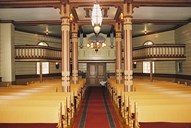 From the interior of the church seen towards the gallery and the porch. The colour scheme is fairly similar to the original colurs. The walls are painted yellow, and the nave has wainscoting. The brown-painted pews were last redecorated in 1994. There is a votive ship model hanging from the ceiling. The organ from 1914 has eight registers, and is built by 'Olsen & Jørgensen'.