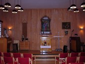 The interior of the 'bedehus': The rostrum, table with a bible, and a large Jesus picture on the wall behind. A cross standing on the floor. Two other pictures: Jesus and Pilate, and Jesus in the garden of Gethsemane. To the left of the rostrum hangs a tapestry made with a tatting technique. This was made by a seaman.