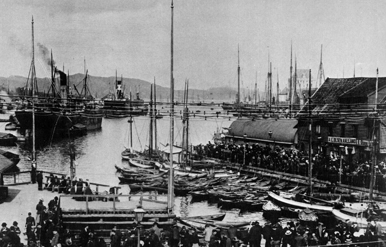 This picture from the Bergen harbour in 1902 shows a number of different cargo vessels, which are in town to sell products, and rowboats, all kinds of big boats, "jakts" and "jekts". Several sloops with gaff sails are docked with the stern to the quayside.<br />
In the front to the left is the triangular quay of Triangelen. This was the best quay, where royalties went ashore. The lack of quay space made steamships anchor in the bay of Vågen and are unloaded and loaded by cargo vessels. At the quay of Zachariasbryggen, there are large crowds around the 60 live-fish tanks, and under the oval roof the steam fire engine is at the ready.<br />
The picture is from F. Beyers kalender, 1981.