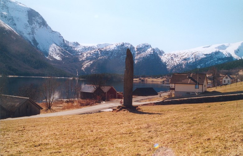 The memorial stone at Ålfoten, erected in 1914 to commemorate soldiers from Ålfoten who served in the war 1807-1814, stands on a cultivated field close to the rural road along the fjord. The road to the right leads up to the church, about 100 metres from the main road.