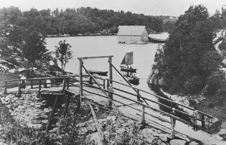 The old bridge across the canal was built with a hoisting device so that the mid section could be opened to let rigged vessels pass through. In the picture we can see rowboats that have just passed through and are on their way into the bay of Gaddevågen. In the background we can see the old Nygård warehouse that is now torn down. The road to the right is the old road leading up to Nygård.