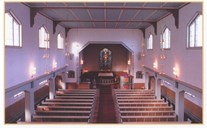 The church nave is light and has a somewhat square look. The church has one row of windows high up on the mid walls, and another row further down on the side-aisle walls. The eye-catcher in the room is the colourful stained-glass painting above the altar. 