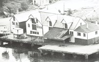 The Hovden Herring Salting Company on the island of Kalvøya. Furthest to the left there used to be an old warehouse, but in all other respects it looks almost as it did in the 1950s. House no. 2 from the left originally contained a factory for producing cod-liver oil on the ground floor, while the first floor was used as dining room and kitchen. Houses nos. 3 and 4 were originally separated houses used by people from the Gulen area in Bremanger. The second floor of house no. 3, as well as the extension furthest to the right, was used for lodging purposes. On the open area there was earlier a storehouse for barrels. To the left on the quay we see a crane and a silo. Under the silo there was a gauge for measuring the weight of herring in hectolitres. From this point the herring was transported to three different conveyor belts. 