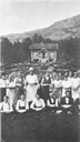 From a summer farm gathering some time in the 1920s. A group of youngsters lined up, holding bowls of porridge. One of the girls is holding a magazine showing a picture of a man. The cabin in the background used to be called "Skrivarhytta" because it was owned by a district recorder. Later on it was named "Fønstelienhytta" after the owners, the Fønstelien family in Måløy.